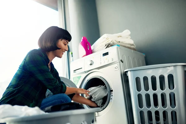 Person loading laundry into a front-loading washing machine in a bright, modern laundry room with towels and detergent on top.