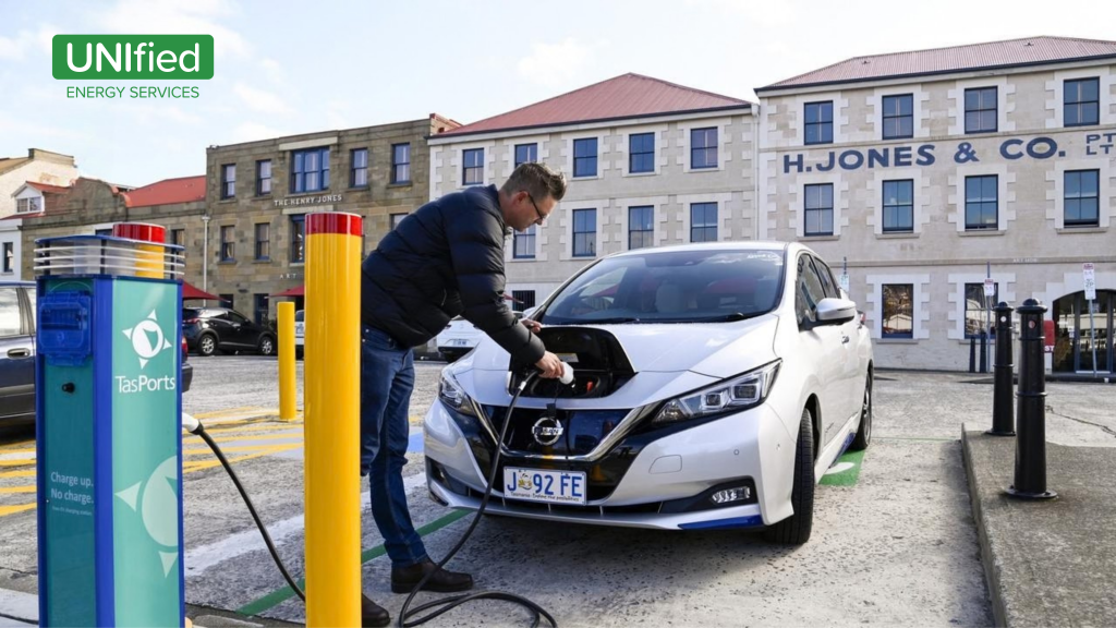 A man charges a white Nissan electric vehicle at a TasPorts public charging station marked “Charge up. No charge,” . Australia fuel prices EV solar batteries